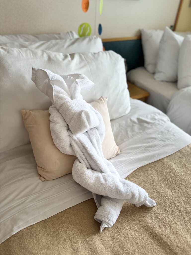 A towel animal designed to look like a sleeping bunny set against a pillow on a cruise ship bed.