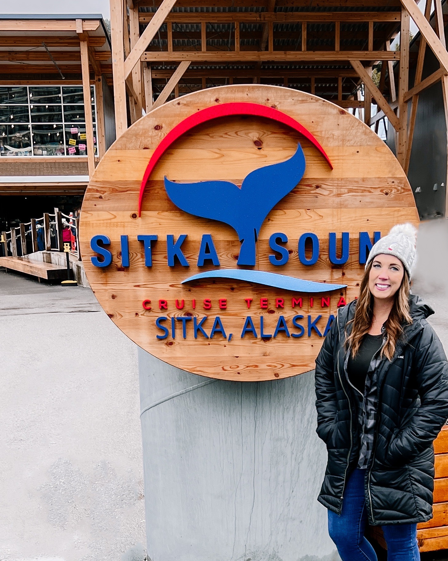 A woman in a coat and winter hat poses in front of the welcome sign at the Sitka cruise port in Sitka, Alaska