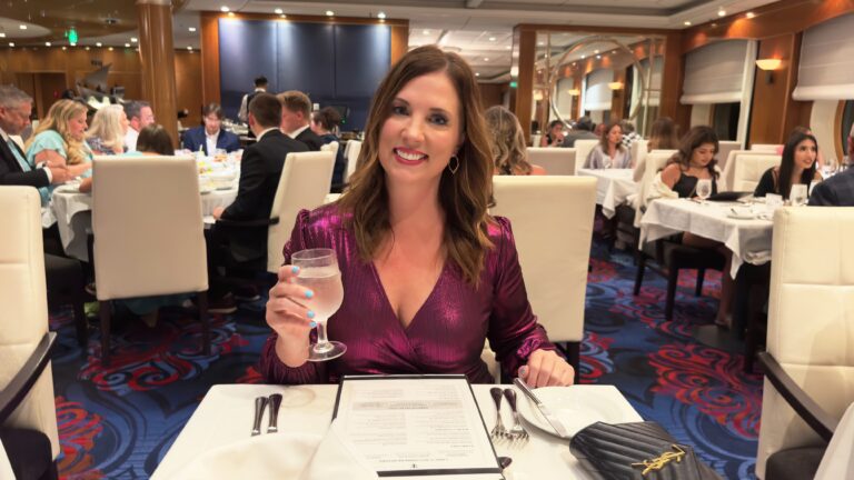 A woman poses with a glass of water in her hand at a table in a cruise ship Main Dining Room