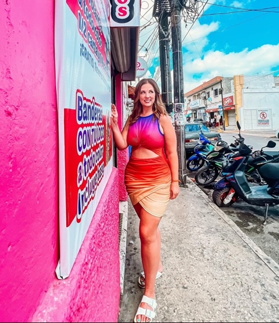 A woman in a colorful dress poses next to a vibrant pink wall with motorcycles parked behind her in downtown Progreso, Mexico