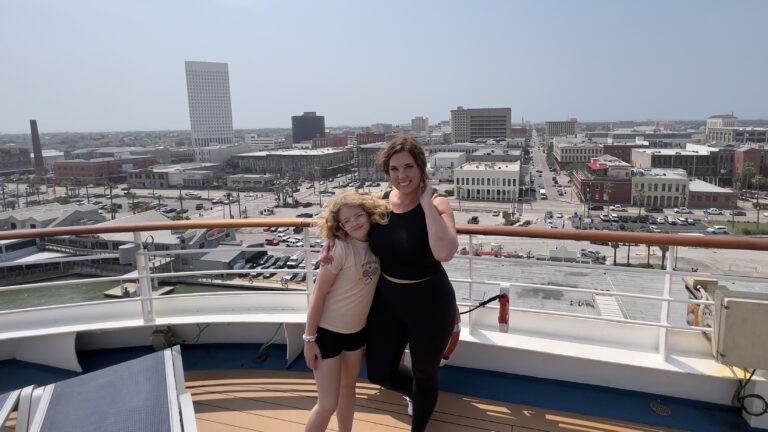 A woman and her daughter pose on a Carnival cruise ship docked in Galveston, Texas, with the Galveston skyline visible behind them.