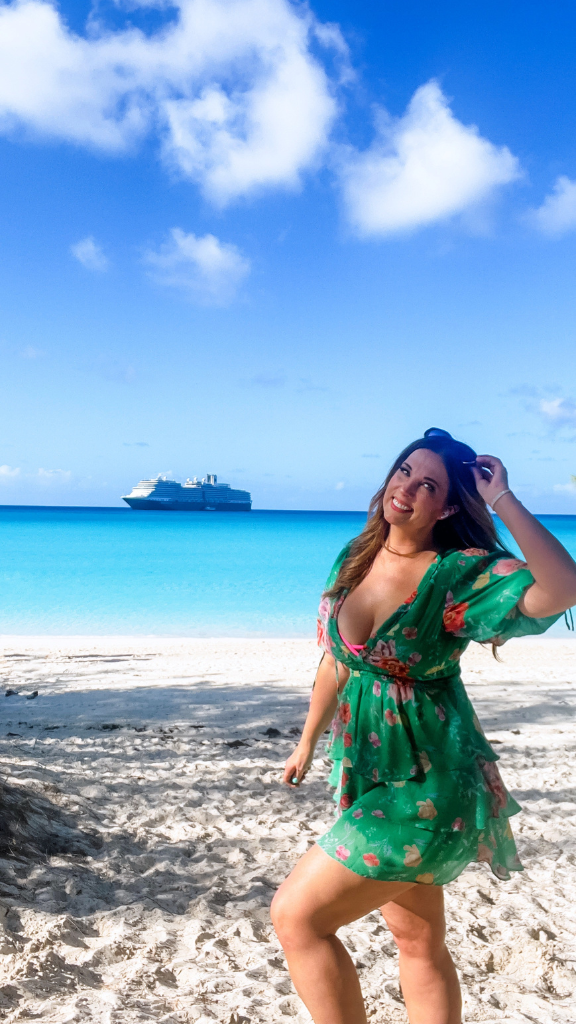 A woman in a green floral dress is standing on a sandy beach with crystal-clear turquoise waters in the background.