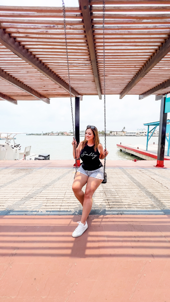 A woman wearing a black tank top and white shorts sits on a swing under a wooden pergola by the waterfront.