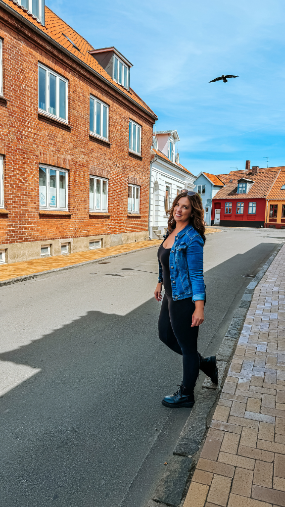 A woman in a denim jacket and black outfit stands on the side of a quiet street lined with historic brick buildings in Visby. 