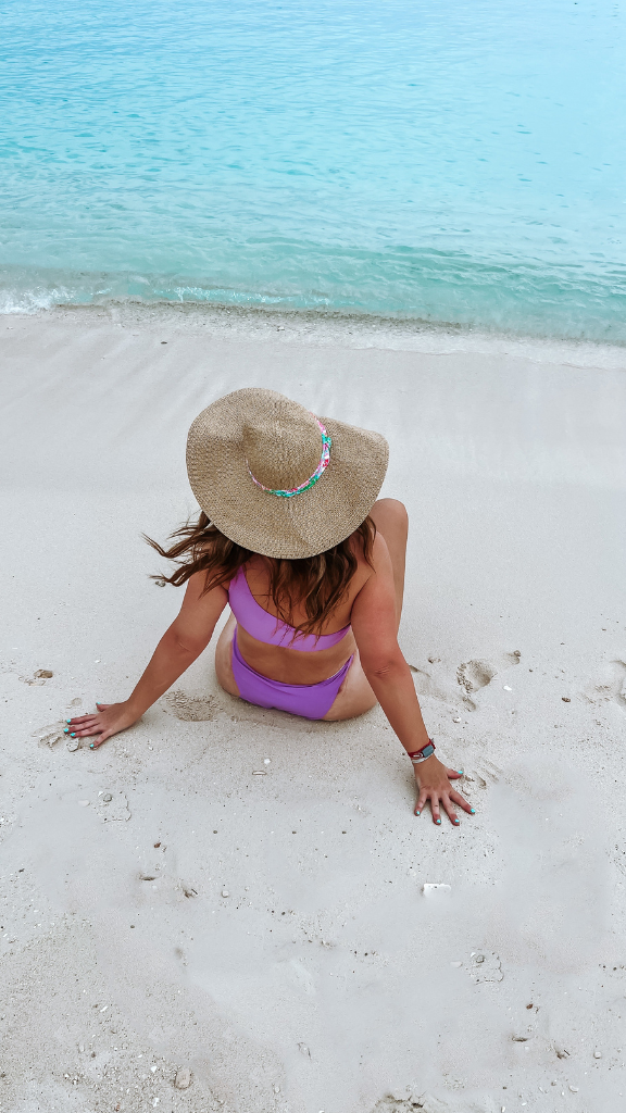 A woman wearing a purple bikini and a wide-brimmed straw hat sits on the sandy beach, facing the turquoise ocean.