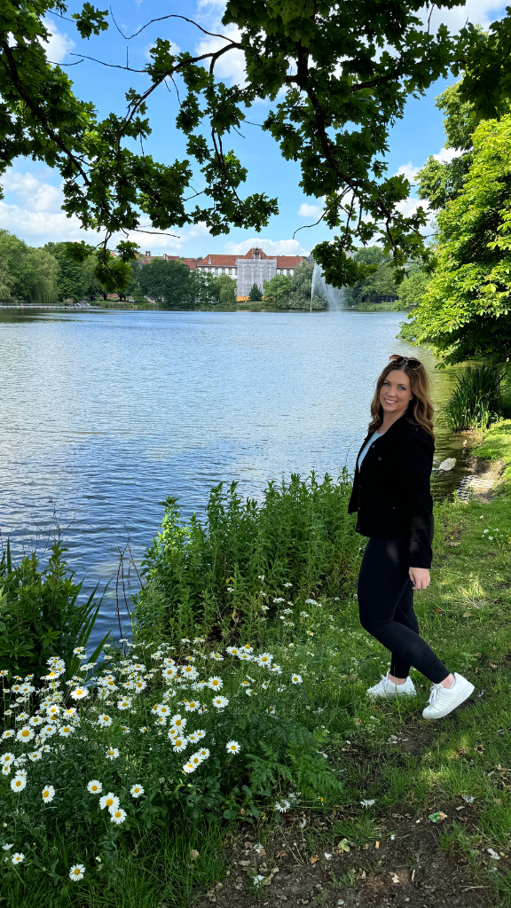 A woman stands by the edge of a serene lake surrounded by greenery and flowers. She is smiling and wearing a black jacket, black pants, and white sneakers.