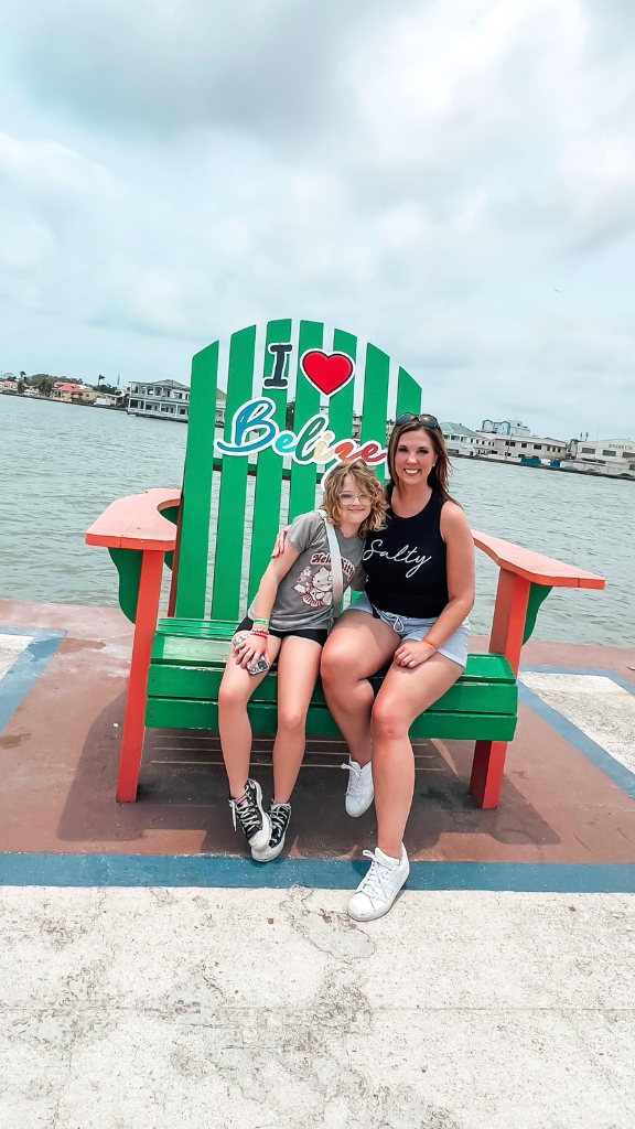 Two women are sitting on a large green Adirondack chair with "I ❤️ Belize" written on it. They are smiling and enjoying their time by the waterfront.