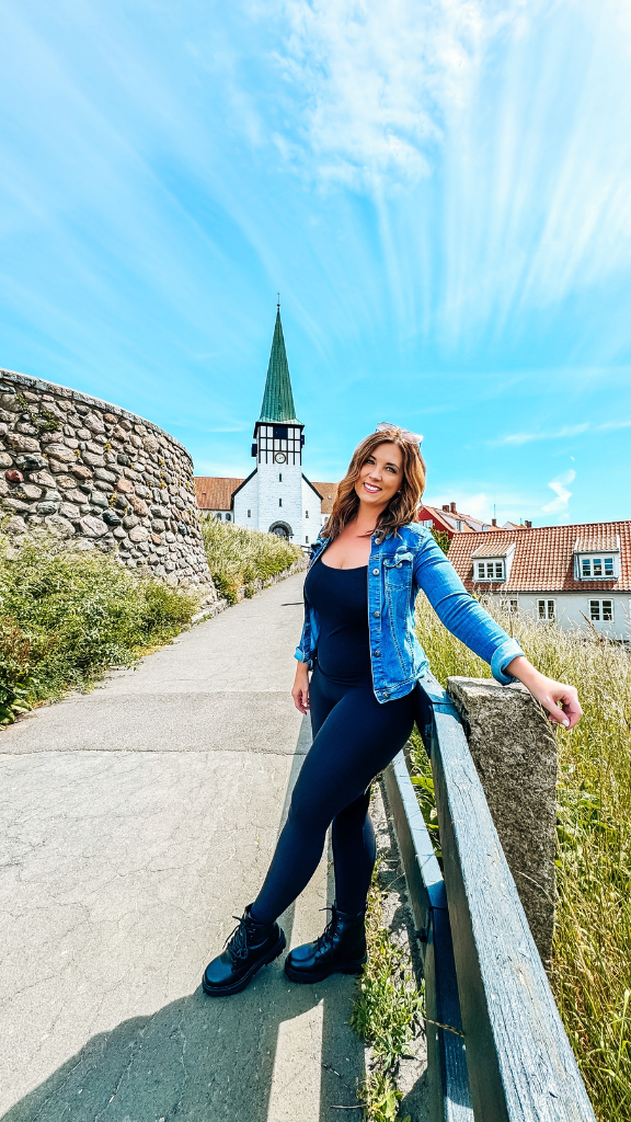 A woman in a denim jacket and black outfit poses on a pathway next to a stone wall in front of a historic church with a green steeple.