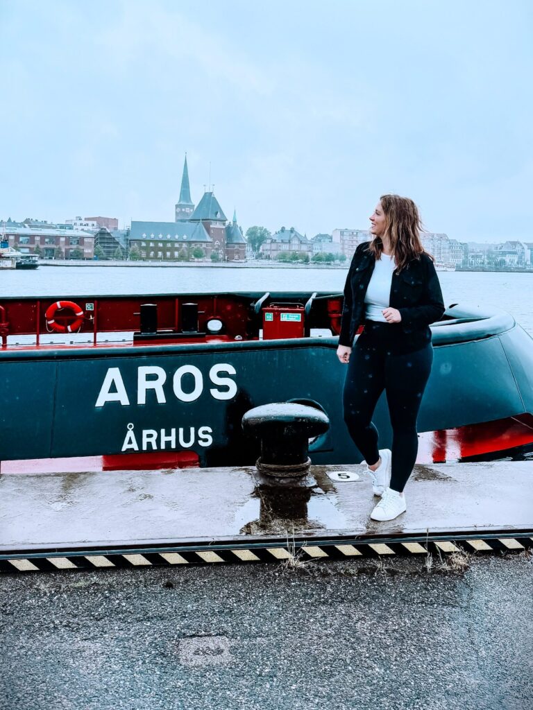 A woman stands in front of a boat labeled "Aros: Arhus" at the cruise pier in Aarhus, Denmark.