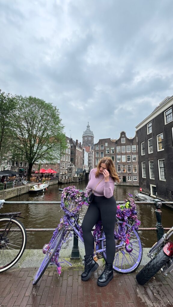 A woman stands with a bike decorated with purple flowers on a bridge over an Amsterdam canal.
