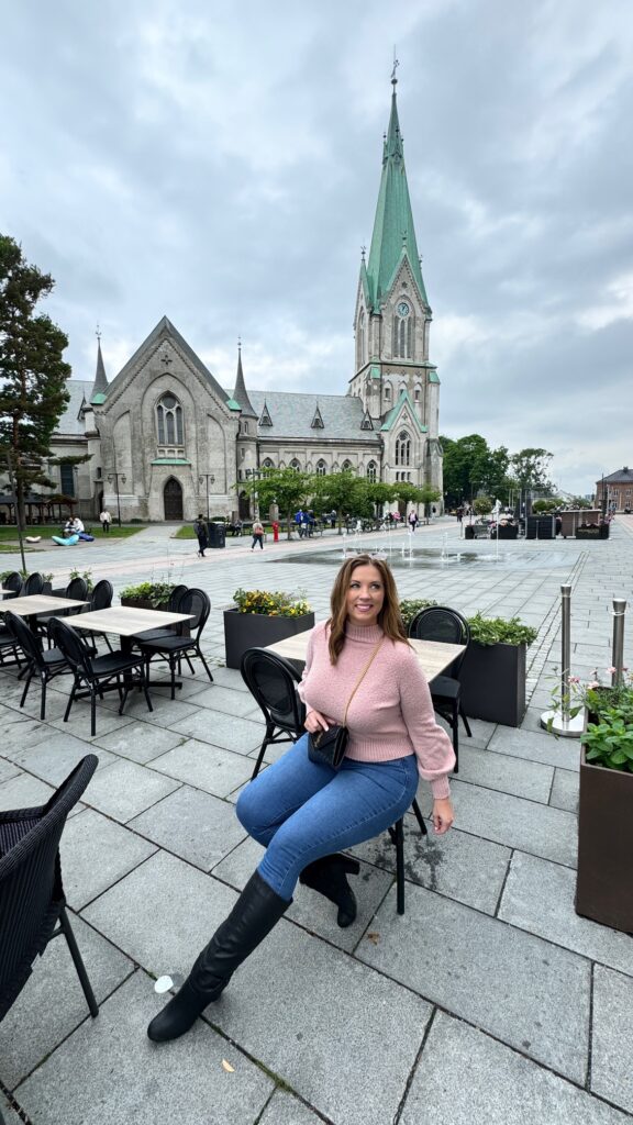 A woman poses in a cafe chair in front of the Kristiansand Cathedral in Norway.