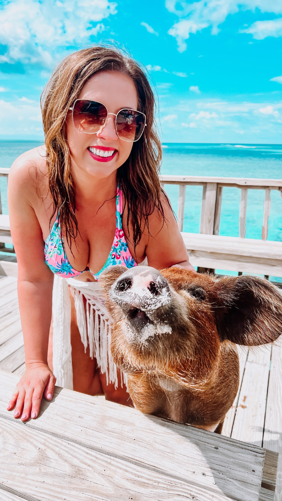 A woman wearing sunglasses and a floral bikini top is smiling and posing with a pig on a wooden deck by the ocean.