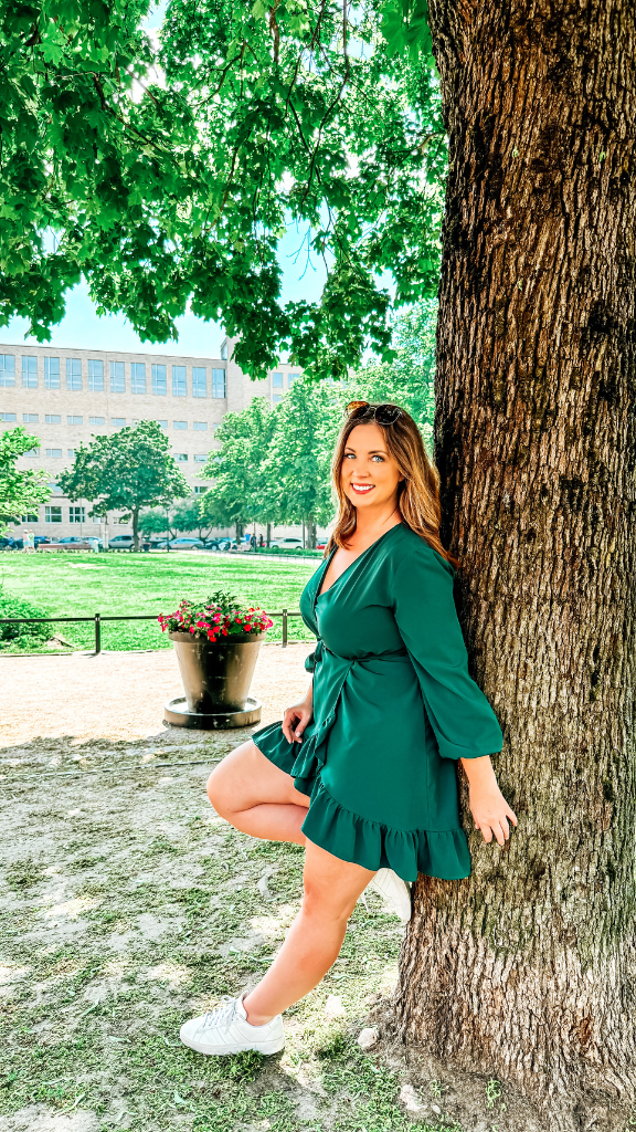 A woman wearing a green dress and white sneakers is leaning against a large tree in a park.