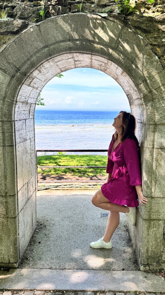 Smiling woman in a bright pink dress leaning against a stone archway, with a scenic view of the sea and a clear blue sky in the background.