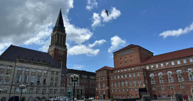 A historic building complex with a prominent clock tower and red brick structures under a blue sky with scattered clouds. A seagull is flying in the sky, adding a touch of movement to the serene scene.