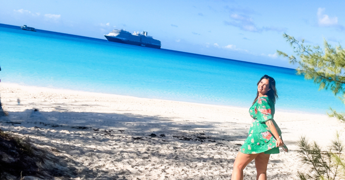 A woman in a green floral dress stands joyfully on a sandy beach with clear turquoise waters in the background.