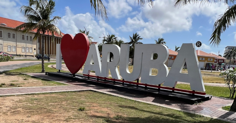 A large sign reading "I ❤️ Aruba" in a park with palm trees and buildings in the background, under a blue sky with scattered clouds.