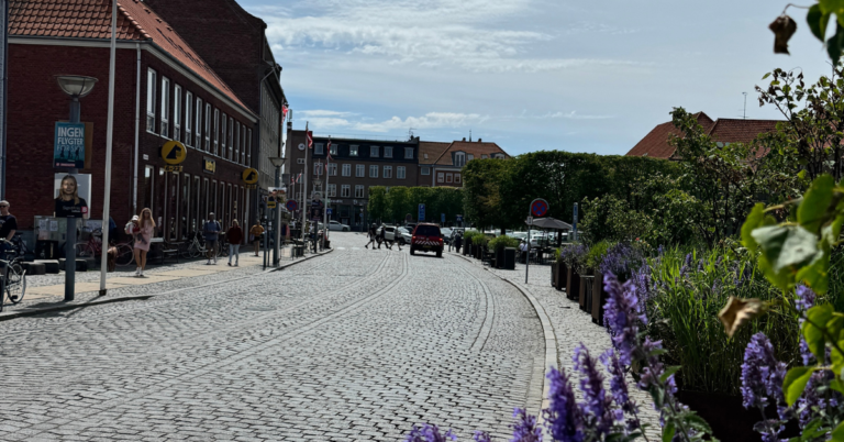 A cobblestone street in a quaint town lined with brick buildings and small shops. People are seen walking and biking along the street.