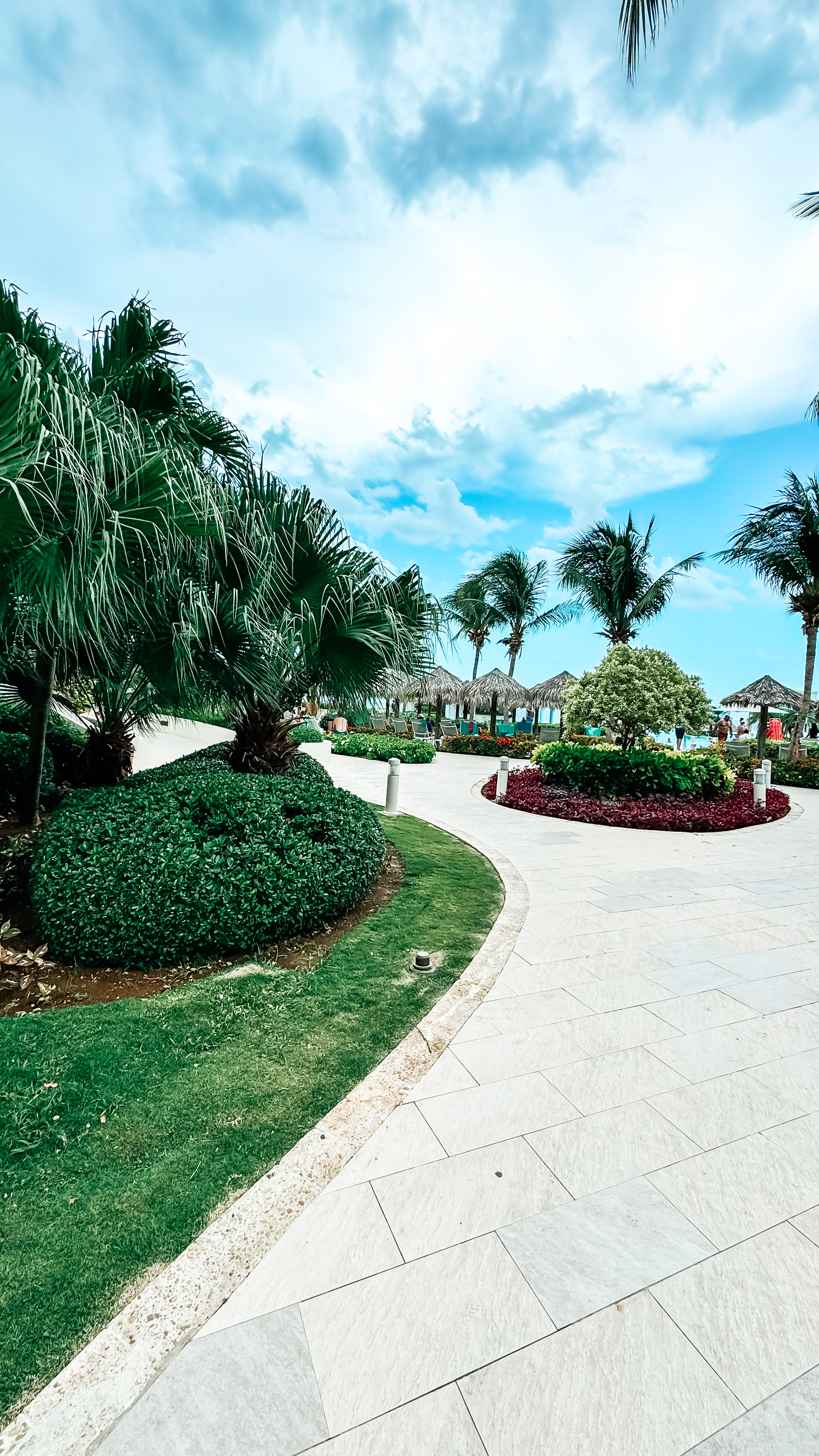 A paved pathway surrounded by manicured lawns, palm trees, and vibrant plants leading towards the beach area under a partly cloudy sky.