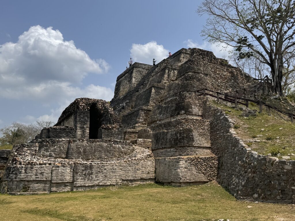 A close-up view of ancient Mayan ruins under a blue sky with a few clouds, showcasing the intricate stone structures and steps. A few people are seen standing on top of the ruins.