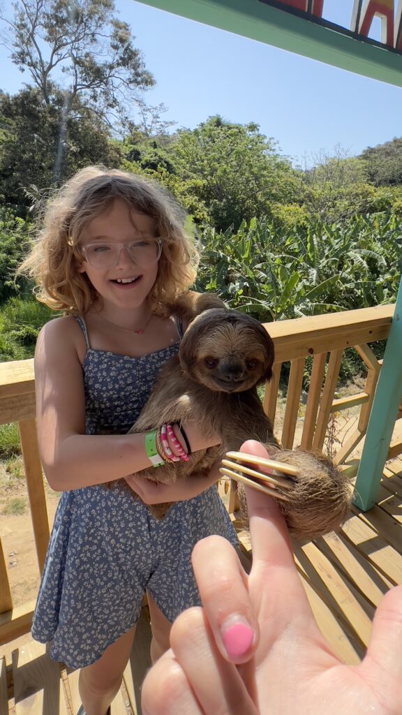 A young girl happily holding a sloth at Manawakie Park, surrounded by lush greenery.