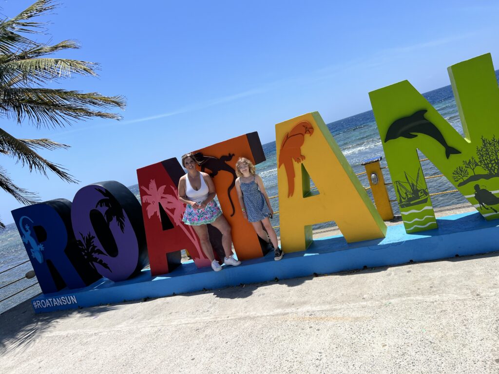 Two people standing next to colorful "Roatan" sign by the beach, with palm trees and the ocean in the background.