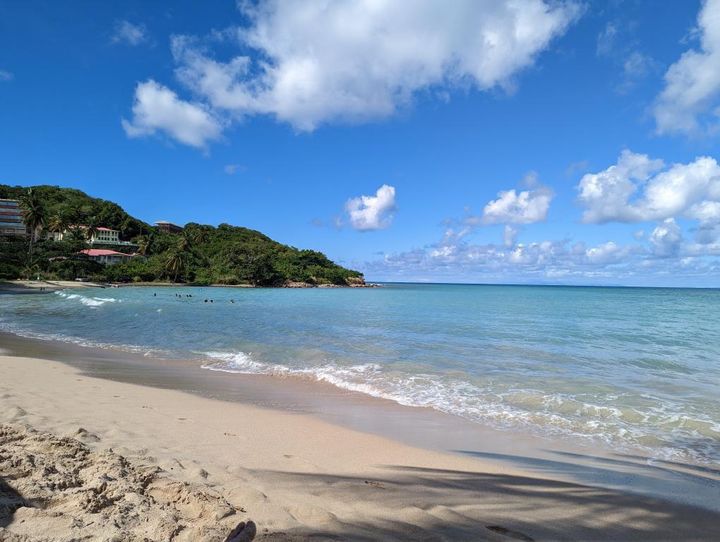 A serene sandy beach with clear blue waters, a few swimmers in the distance, and lush green hills in the background under a partly cloudy sky.