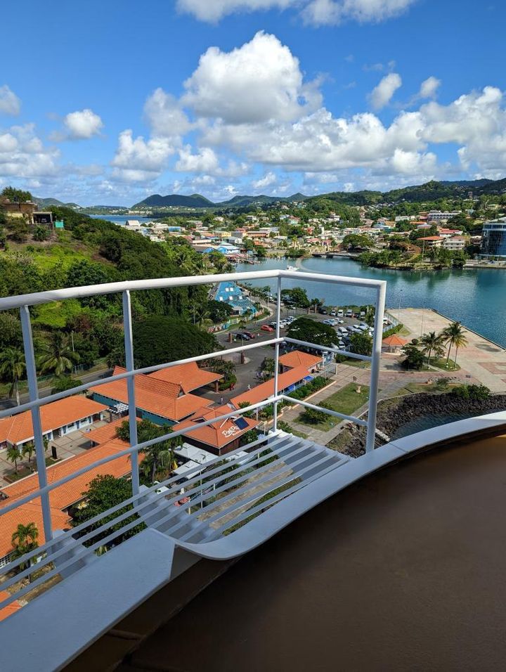 A panoramic view from a balcony overlooking a coastal town with red-roofed buildings, green hills, and a clear blue bay under a partly cloudy sky.